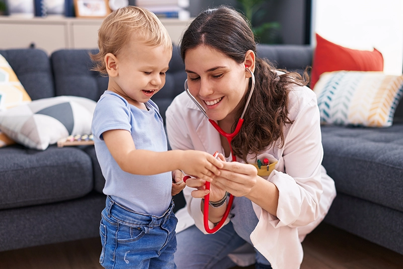 A pediatric nurse in blue scrubs showing a stethoscope to a curious young child at home, demonstrating a friendly approach to pediatric care at home.