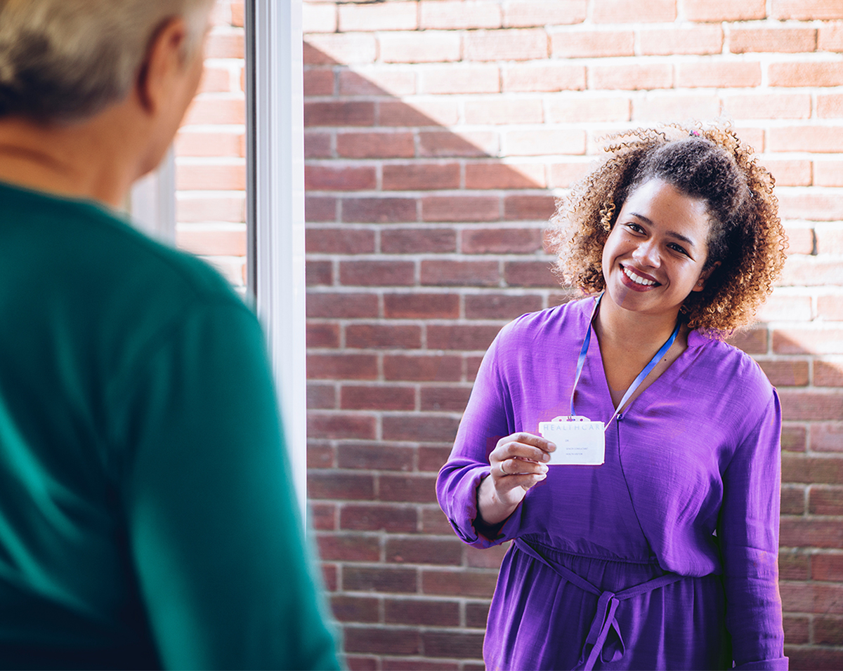 A healthcare worker stands at the front door of a senior woman's home, displaying her badge with a warm, welcoming smile. She is there to provide personal care services, emphasizing the comfort and security of receiving healthcare in one's own home.