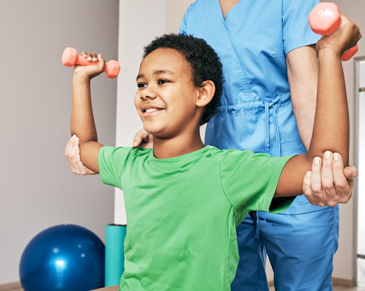 A specially trained physical therapist assists a child with strength training at home, using a small weights to enhance muscle development. The therapist provides close, compassionate support during the exercise session in a safe and comfortable home environment.